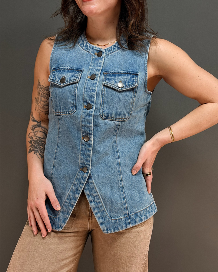Woman wearing sleeveless denim vest and brown pants against a gray background