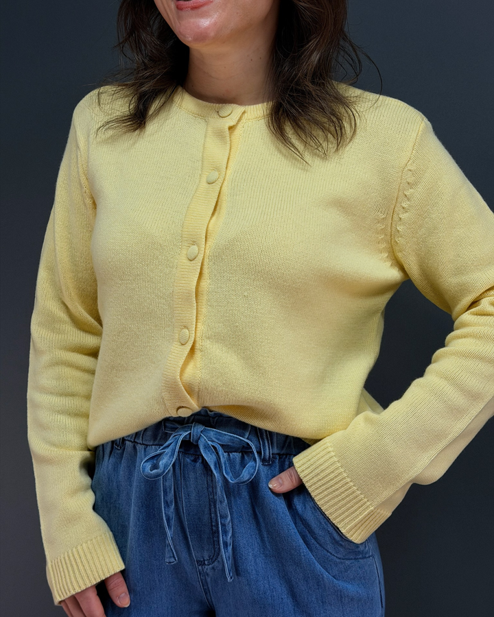 woman wearing yellow button down cardigan with blue tie waist pants in front of a gray background