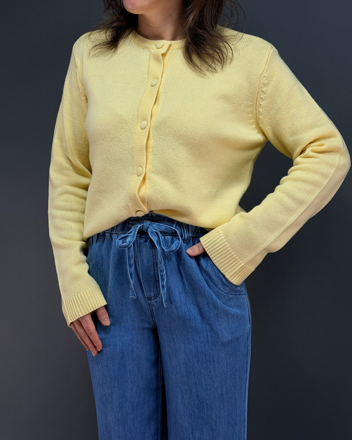 woman wearing yellow button down cardigan with blue tie waist pants in front of a gray background