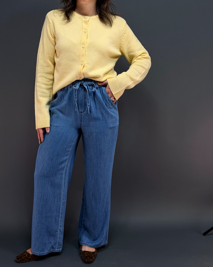 woman wearing yellow button down cardigan with blue tie waist pants in front of a gray background