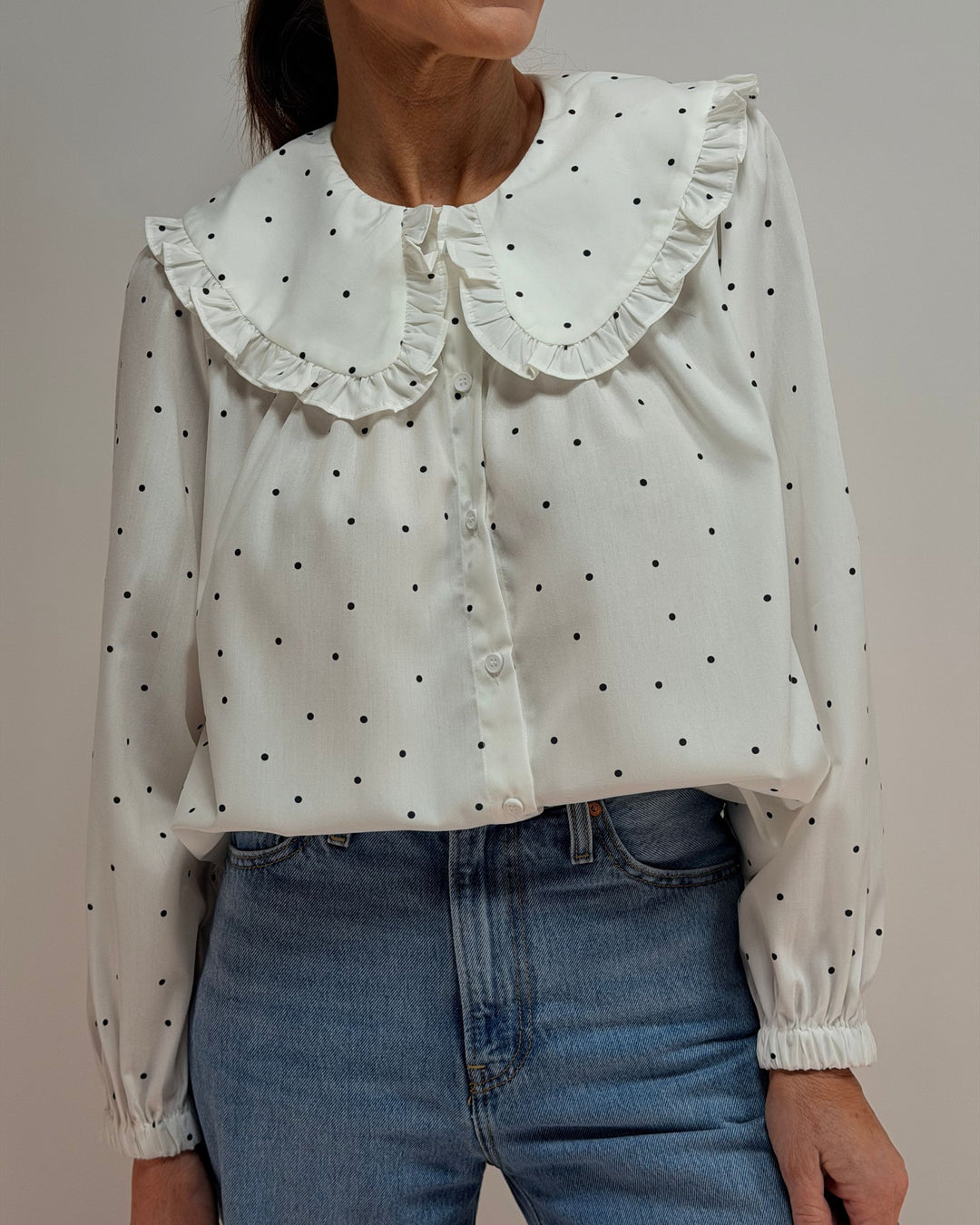 White blouse with black polka dots and ruffled collar worn by a person, against a neutral background.