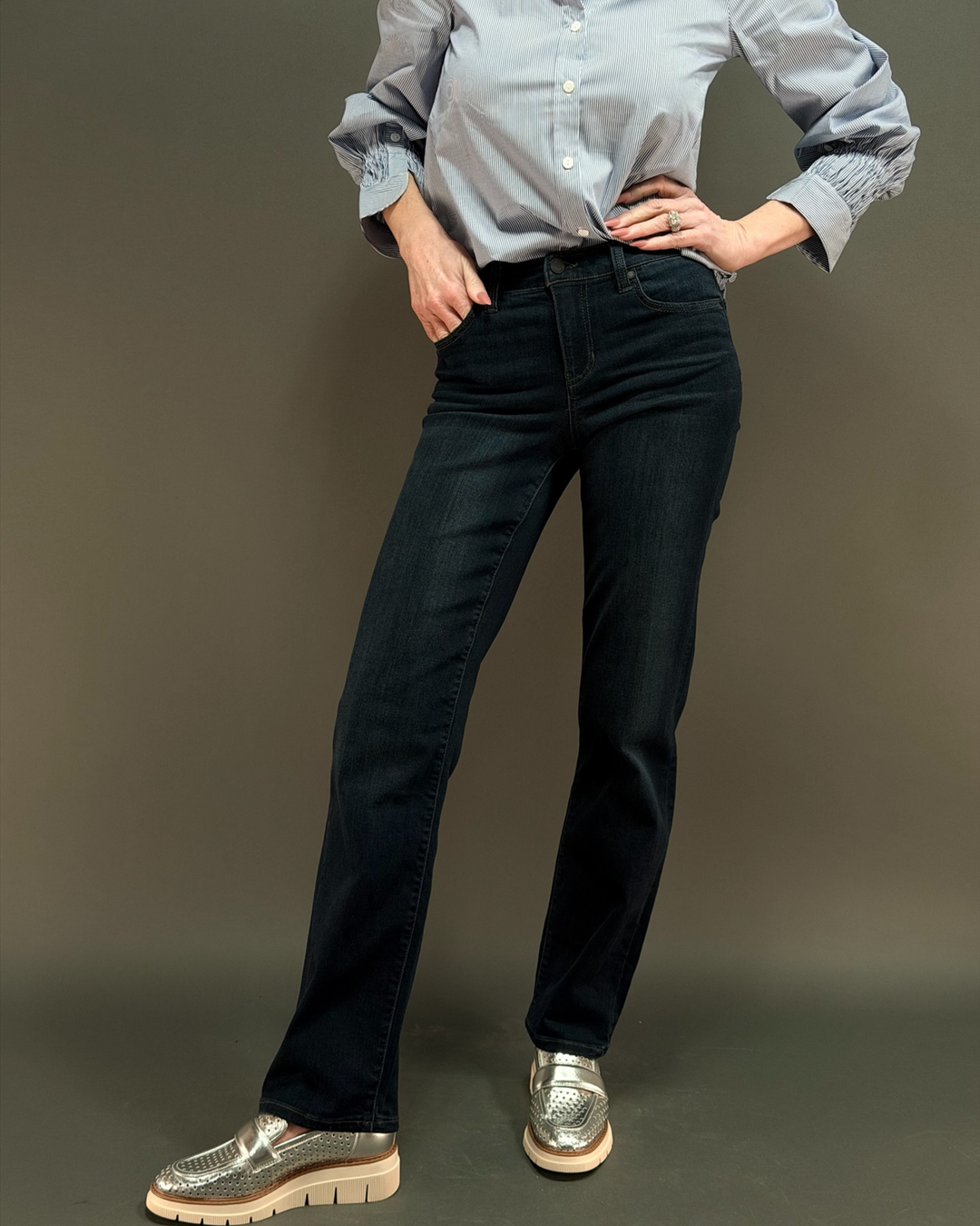 woman wearing dark wash jeans with a blue striped button down in front of a gray background