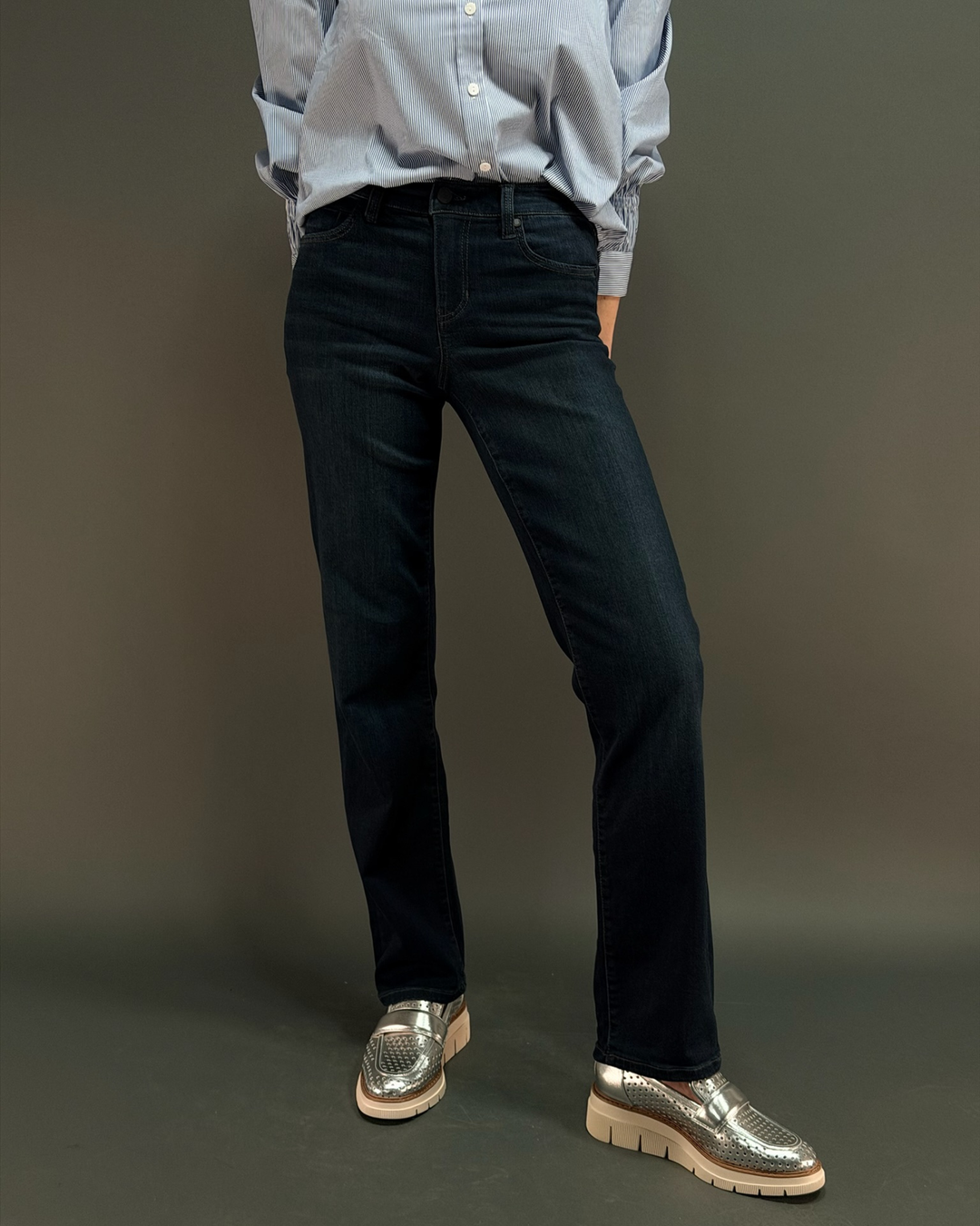 woman wearing dark wash jeans with a blue striped button down in front of a gray background